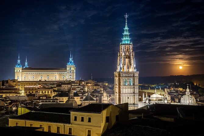 Toledo floodlit after dark, the Alcázar to the left and the Cathedral's spire to the right under a rising moon